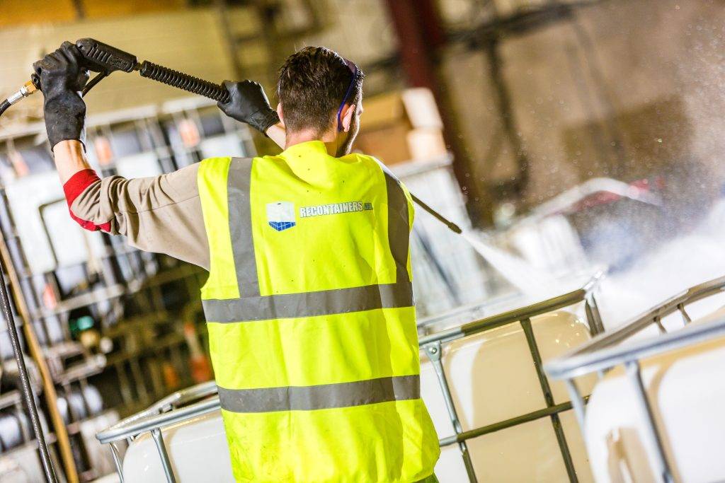 Worker in high-visibility vest pressure washing IBC containers in a warehouse setting for reconditioning.