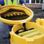 Worker in a yellow vest pours dark liquid from a yellow drum tray into another container, ensuring spill control.