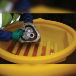 The image shows a person handling a mechanical part in a yellow containment tray with liquid inside.